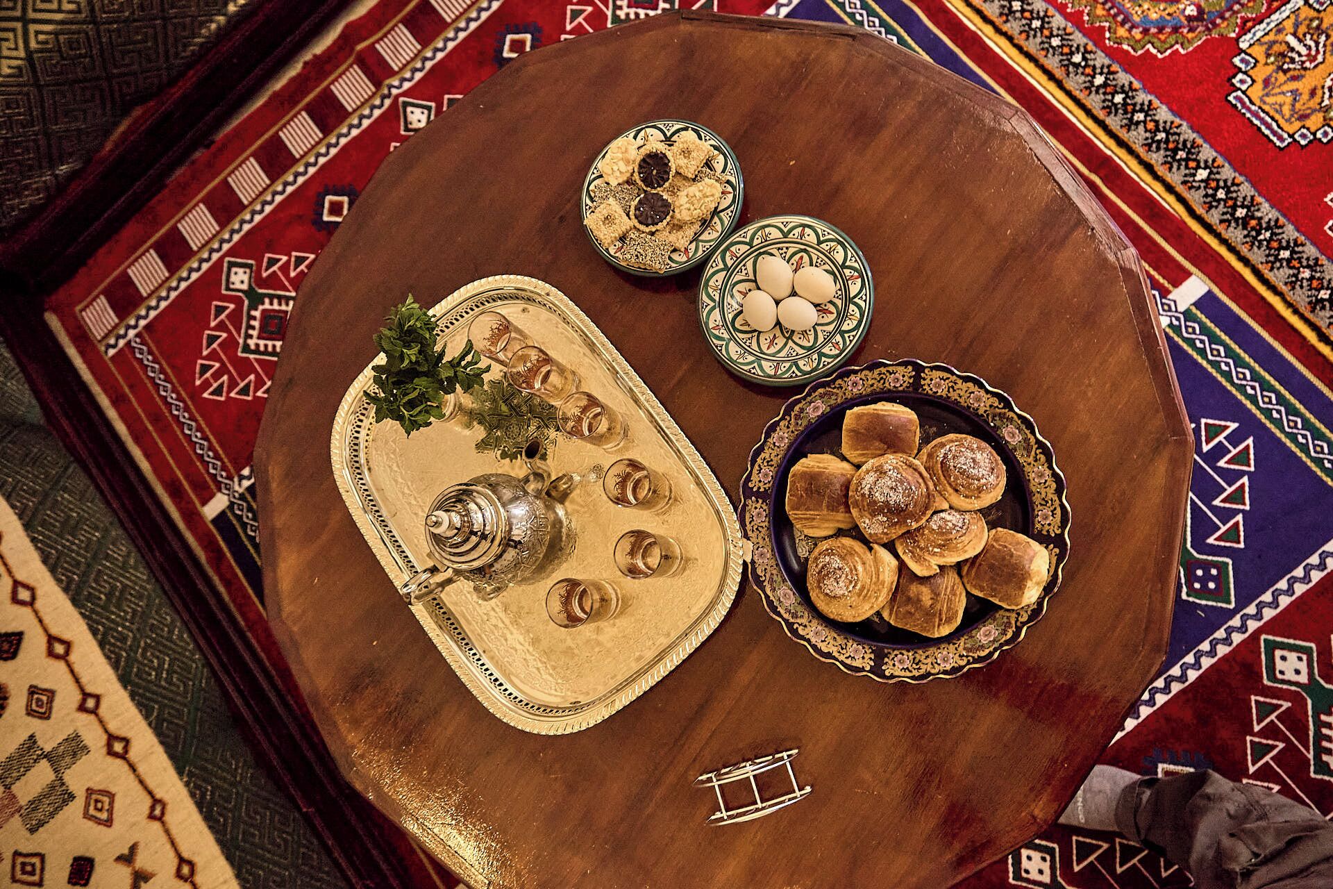 Una mesa redonda de madera con una bandeja que contiene una tetera, vasos y una pequeña planta. También hay platos con galletas y pasteles.