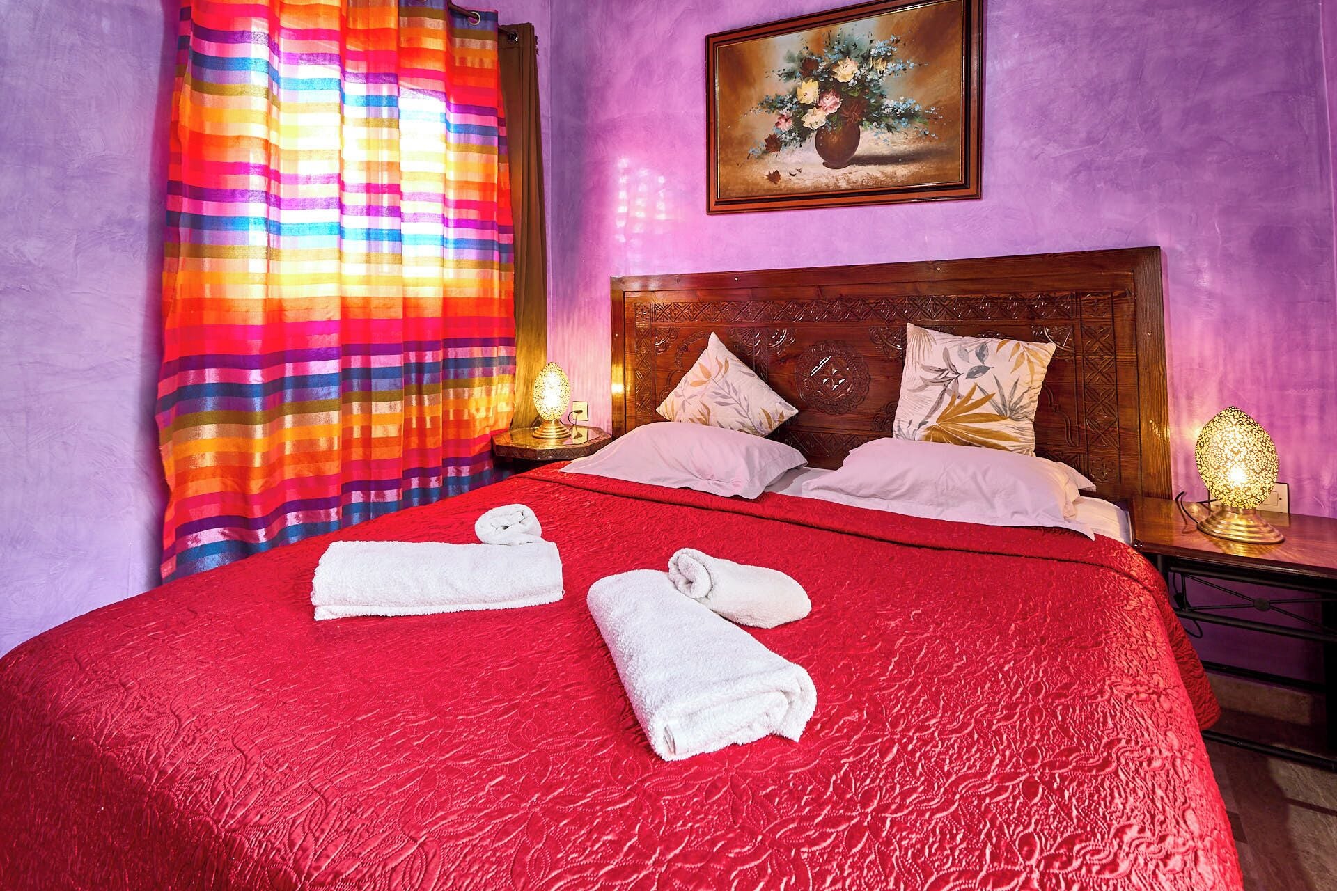 Una cama con una colcha roja, dos almohadas y dos toallas enrolladas.