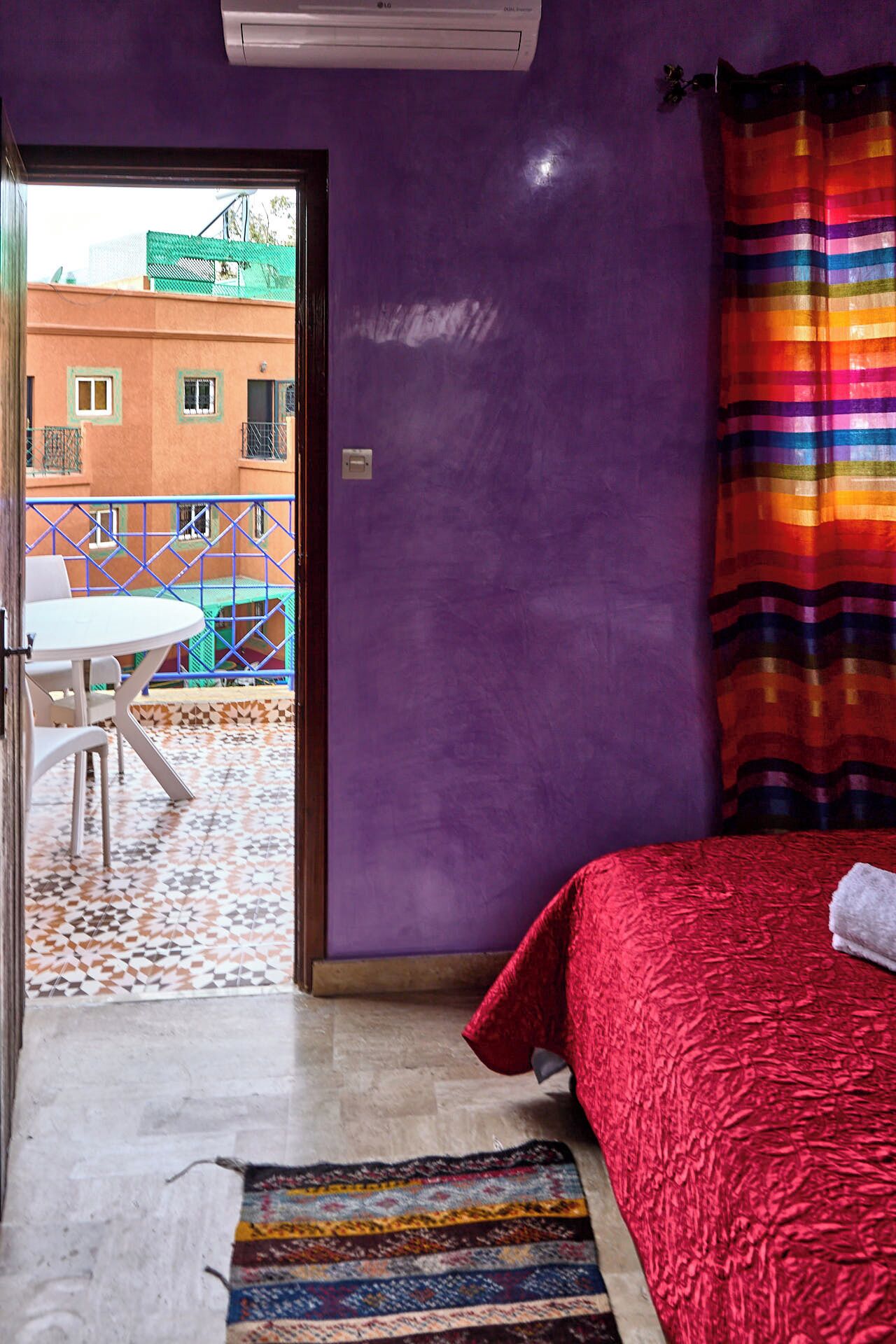 A bedroom with a purple wall, a red bedspread, and a view of a building through an open door.