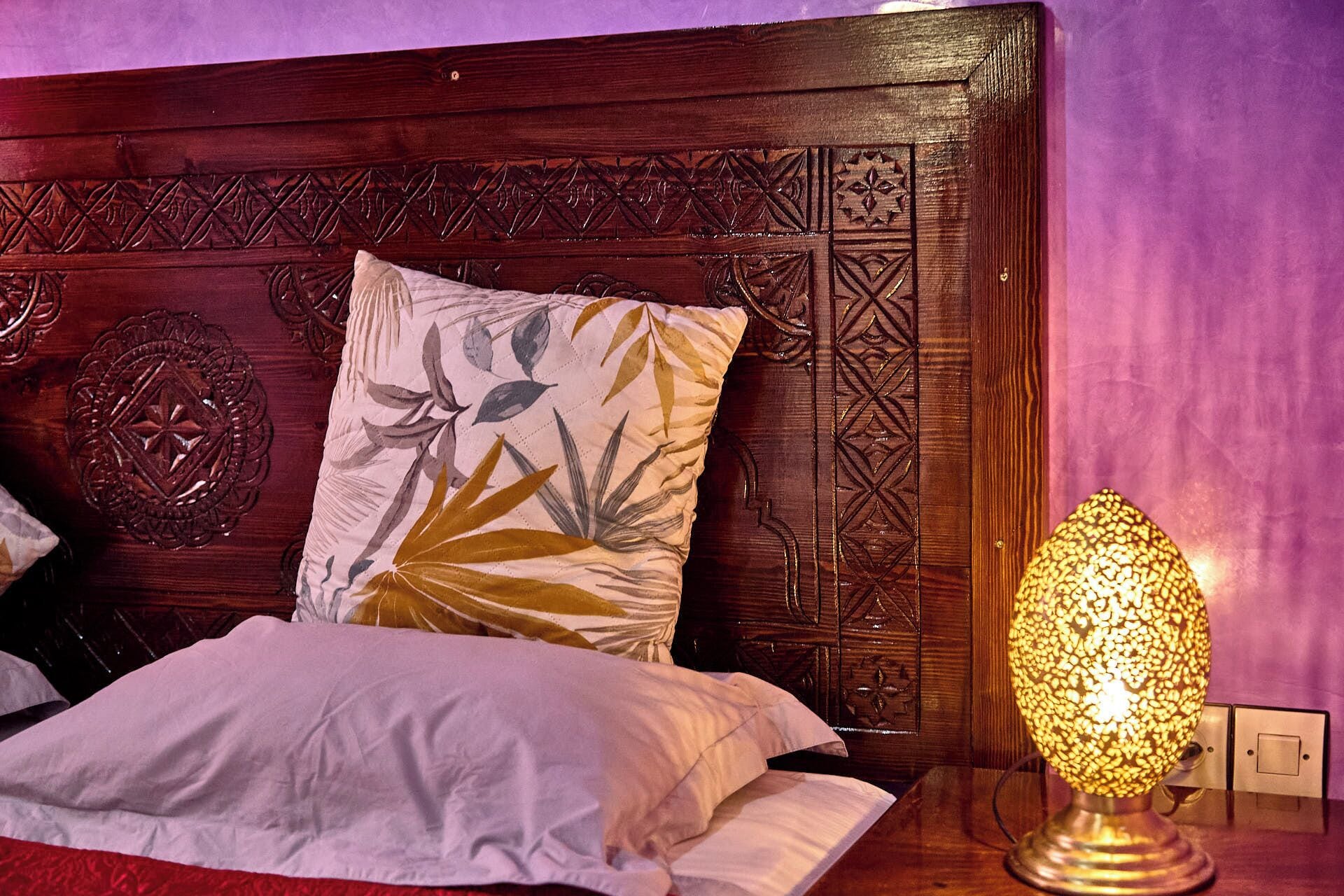 Una cama con un cojín floral, un cabecero de madera con intrincados grabados y una lámpara de mesa con pantalla texturizada.