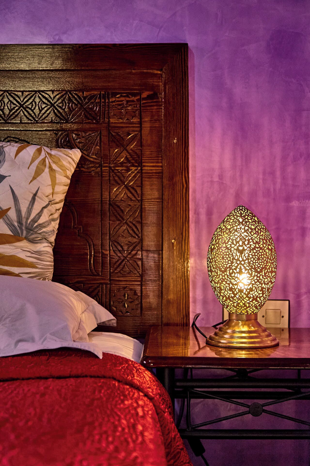 A bed with a red cover, a pillow with a leaf pattern, a wooden headboard with intricate carvings, and a table with a patterned lamp.