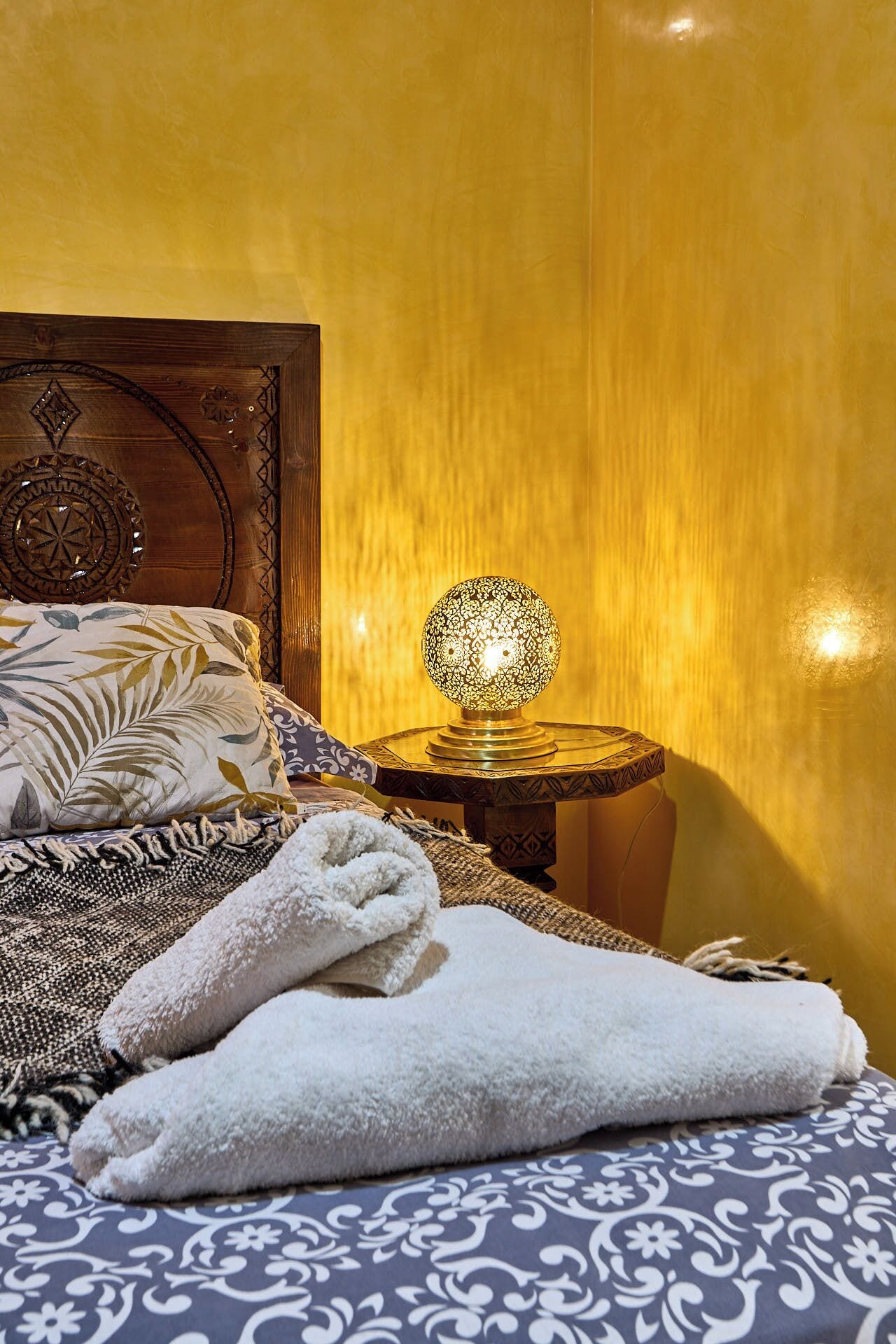 A neatly made bed with a patterned bedspread, a headboard with a decorative design, a bedside lamp with a patterned lampshade, and a small round table with a patterned tablecloth.