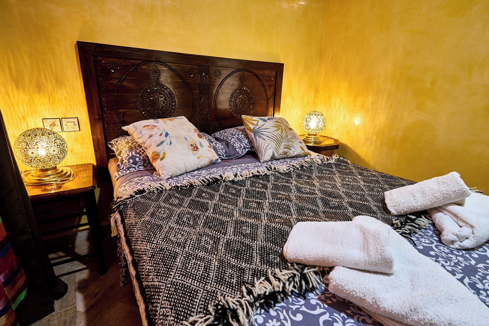 A neatly made bed with a patterned blanket and multiple pillows, a bedside lamp, and a small table with a decorative item.