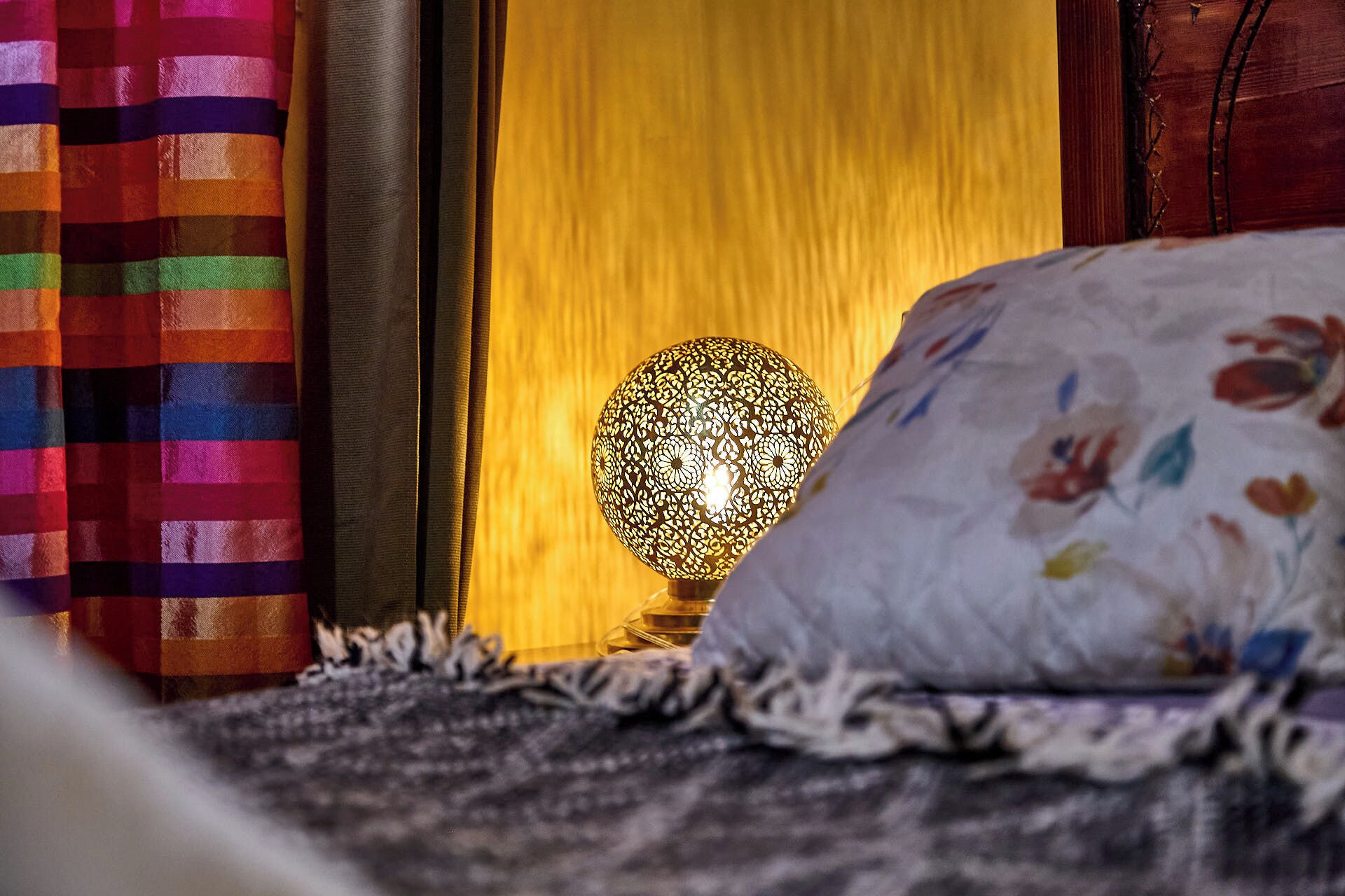 A bed with a floral pillow, a striped blanket, and a lamp with a patterned shade.