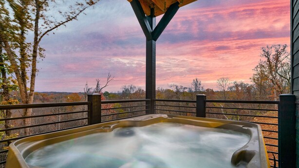Een hottub met een houten dak en metalen reling, omringd door bomen en een uitzicht op de ondergaande zon.