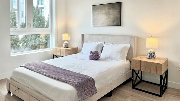 Quarto moderno com uma cama grande, duas mesas de cabeceira e uma janela com vista para edifícios e árvores.