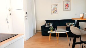 Ein modernes Wohnzimmer mit einem dunkelblauen Sofa, einem kleinen runden Tisch und einem Laptop. Zwei gerahmte Kunstwerke hängen an der Wand und eine Tür mit Schlüsselsperre ist zu sehen.