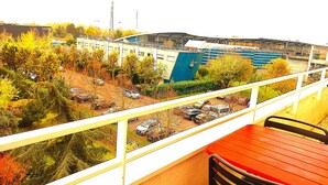 Ein Balkon mit einem roten Tisch und schwarzen Stühlen, der auf einen Parkplatz mit mehreren Autos und ein Gebäude mit blauem Teil blickt.