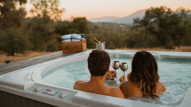 Um casal em uma banheira de hidromassagem, com vista para árvores e montanhas ao fundo.