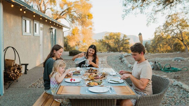 Una familia comiendo al aire libre en una mesa con vistas a una casa y árboles.