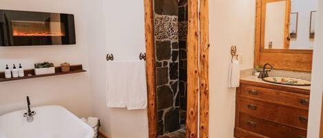 Een badkamer met een douche van natuursteen, houten balken, een wit wastafelmeubel en een spiegel met houten lijst.