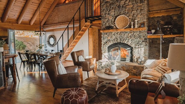 A cozy living room with a stone fireplace, leather furniture, and a wooden staircase. - Scenic View Timber Lodge Hottub by Casa Oso (Mariposa)