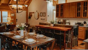 A rustic dining area with a wooden table set for six, surrounded by chairs. A chandelier hangs from the ceiling, and a kitchen with wooden cabinets is visible in the background. - Scenic View Timber Lodge Hottub by Casa Oso (Mariposa)