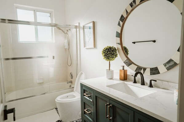 Un baño moderno con cabina de ducha de vidrio, inodoro blanco, lavamanos con un espejo grande y una planta en maceta.