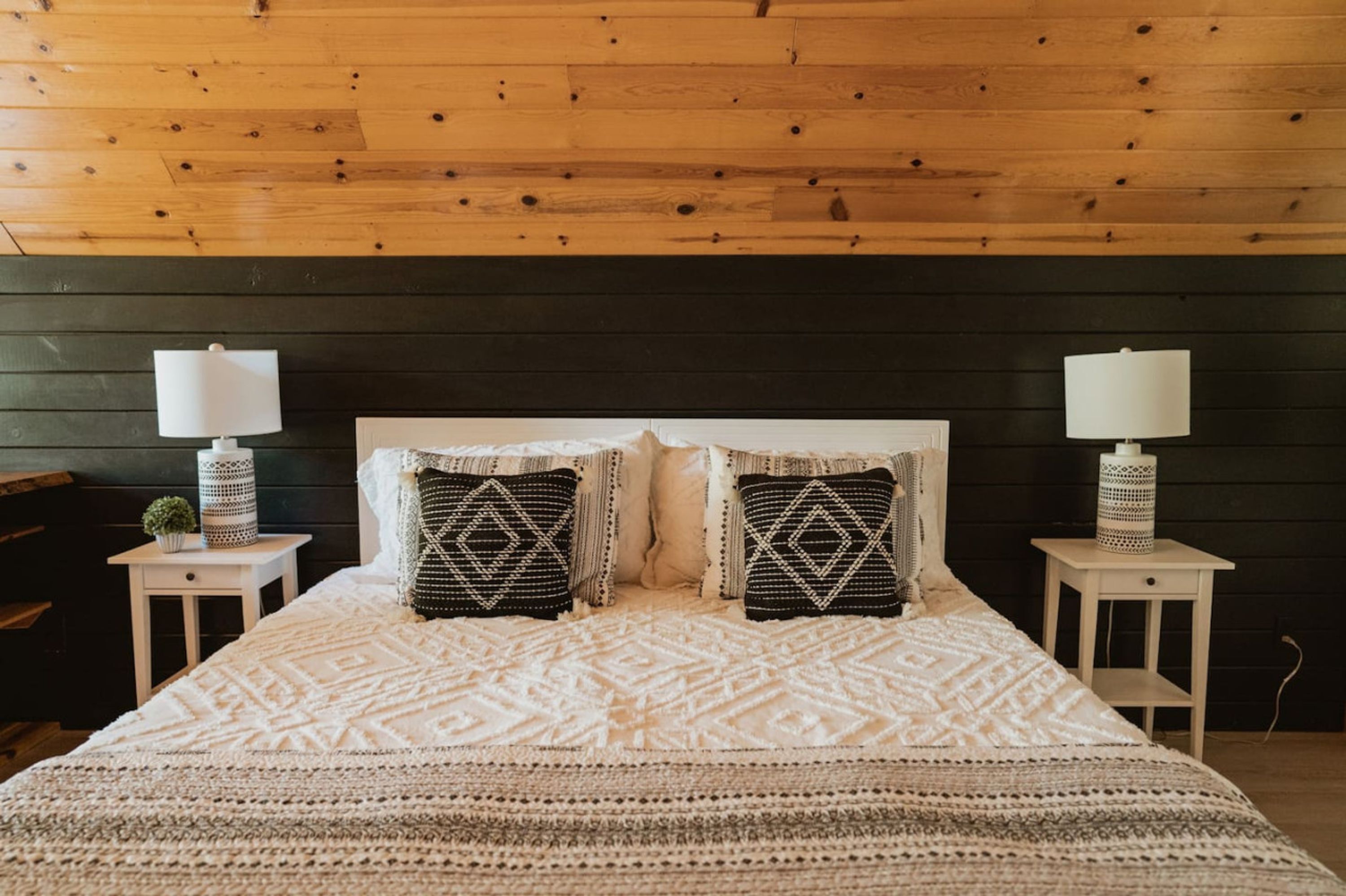 Un dormitorio con techo de madera, una cama con ropa de cama estampada, dos mesitas de noche con lámparas y una planta pequeña en una de ellas.