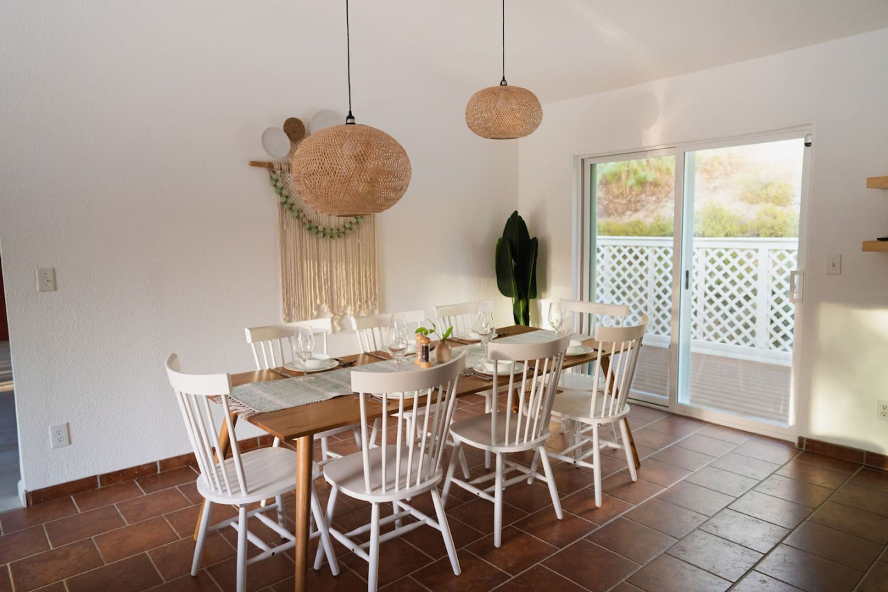 A dining area with a wooden table and white chairs, a macrame wall hanging, and a large window with a view of greenery.