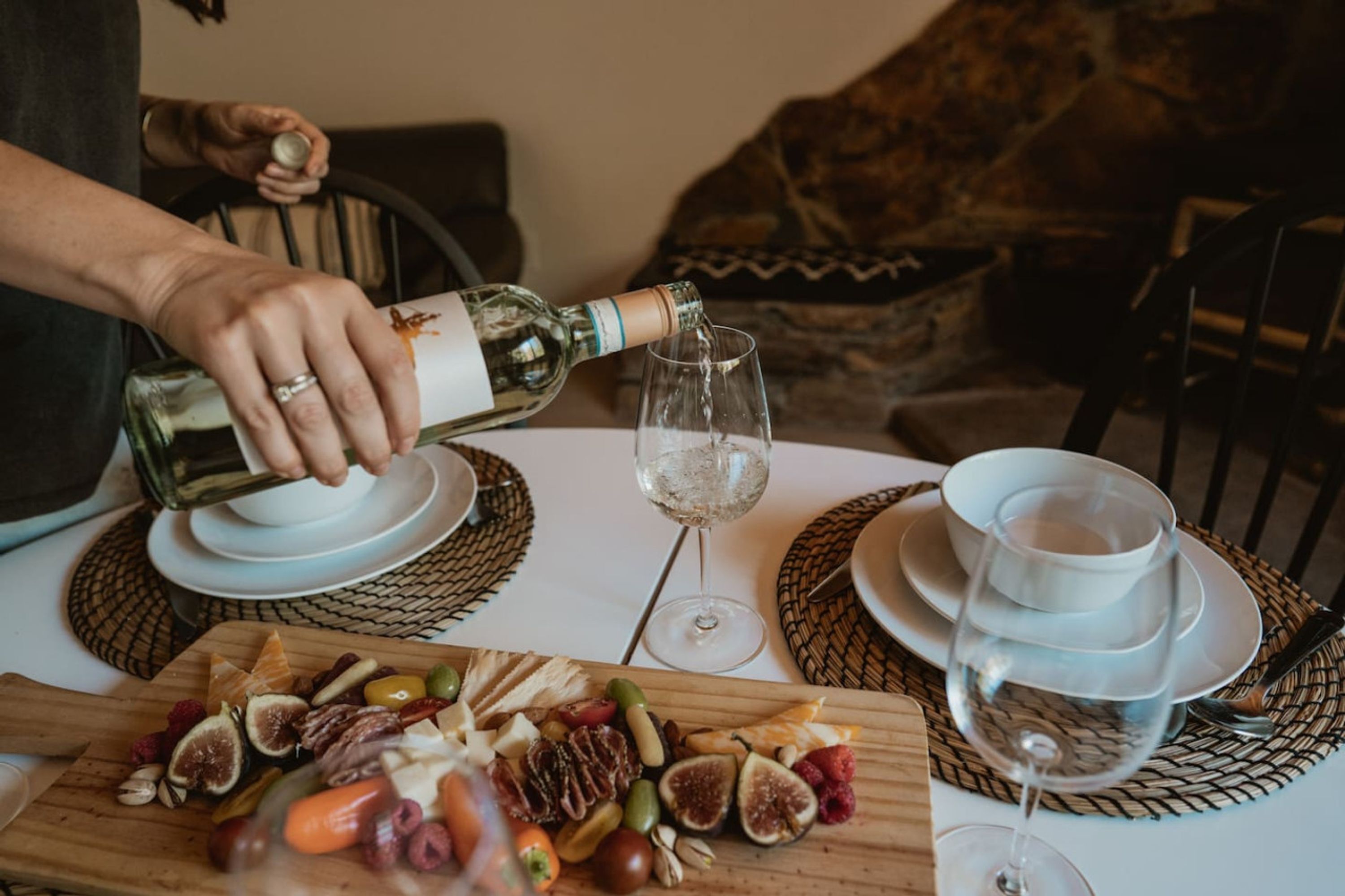 Una persona sirviendo vino en una copa, una tabla de madera con varios quesos y frutas, y un mantel blanco.