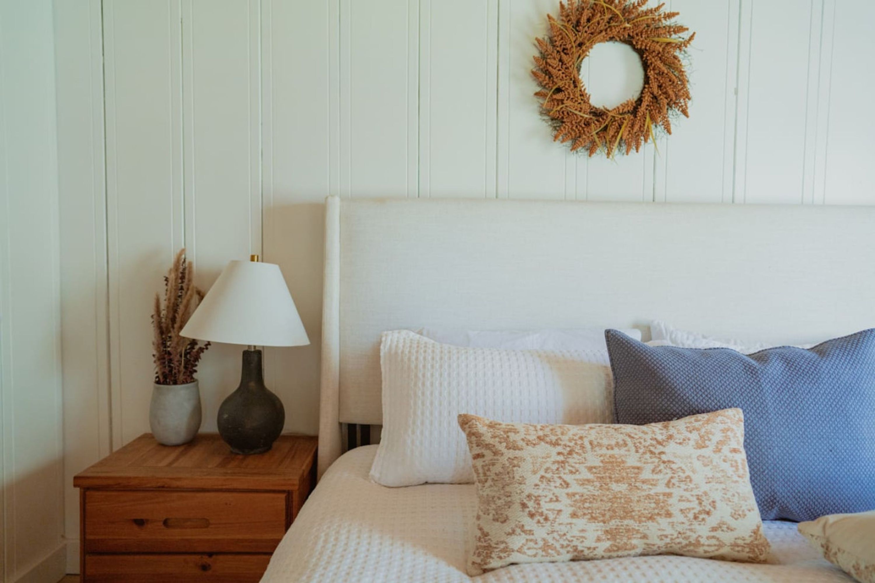Een opgemaakt bed met witte en gemêleerde kussens, een nachtkastje met een lamp en een vaas met gedroogde planten, en een decoratieve krans aan de muur.