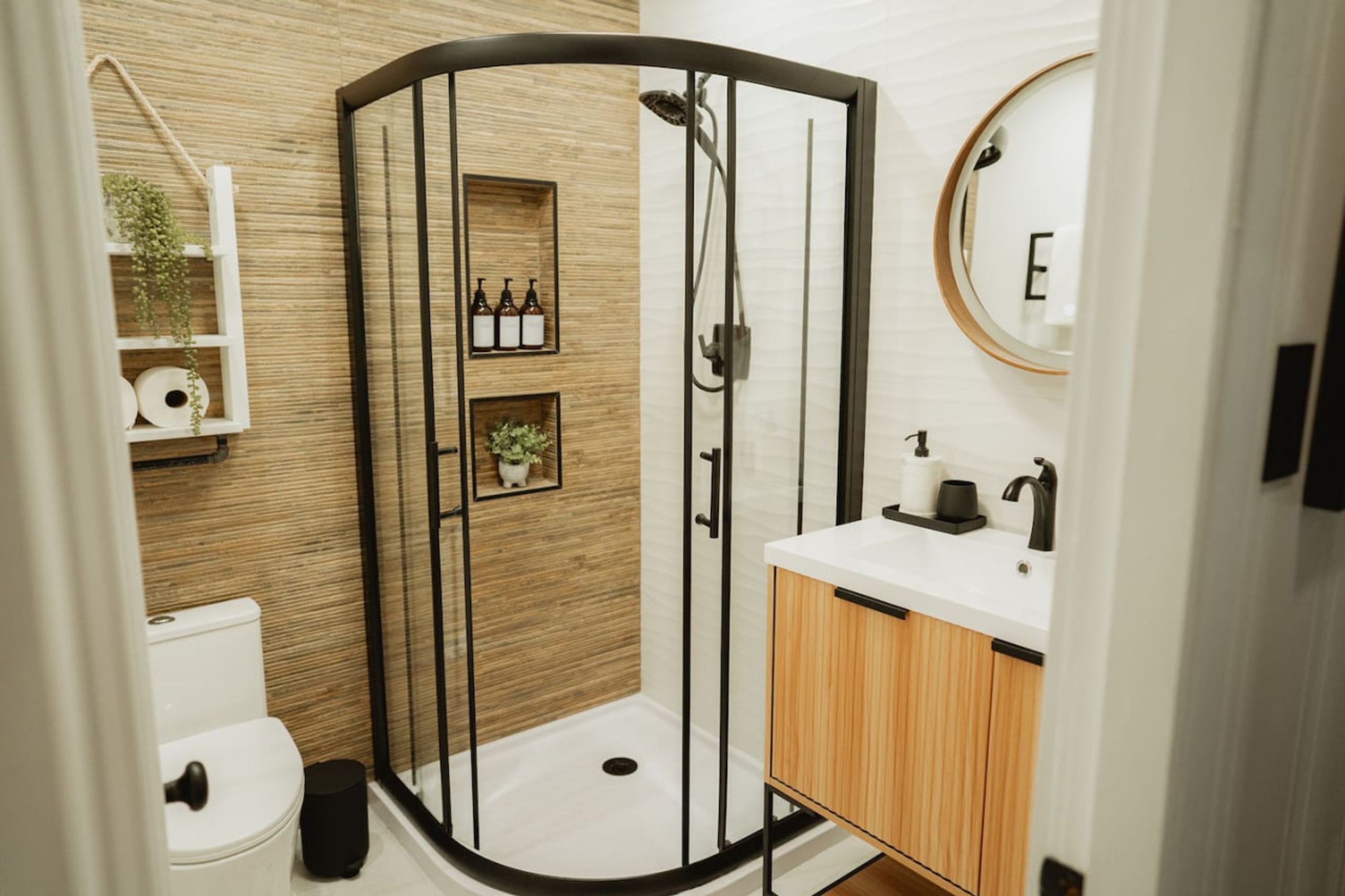 Uma casa de banho moderna com chuveiro envolto em vidro, sanita, lavatório com armário de madeira e prateleira com artigos de higiene pessoal.