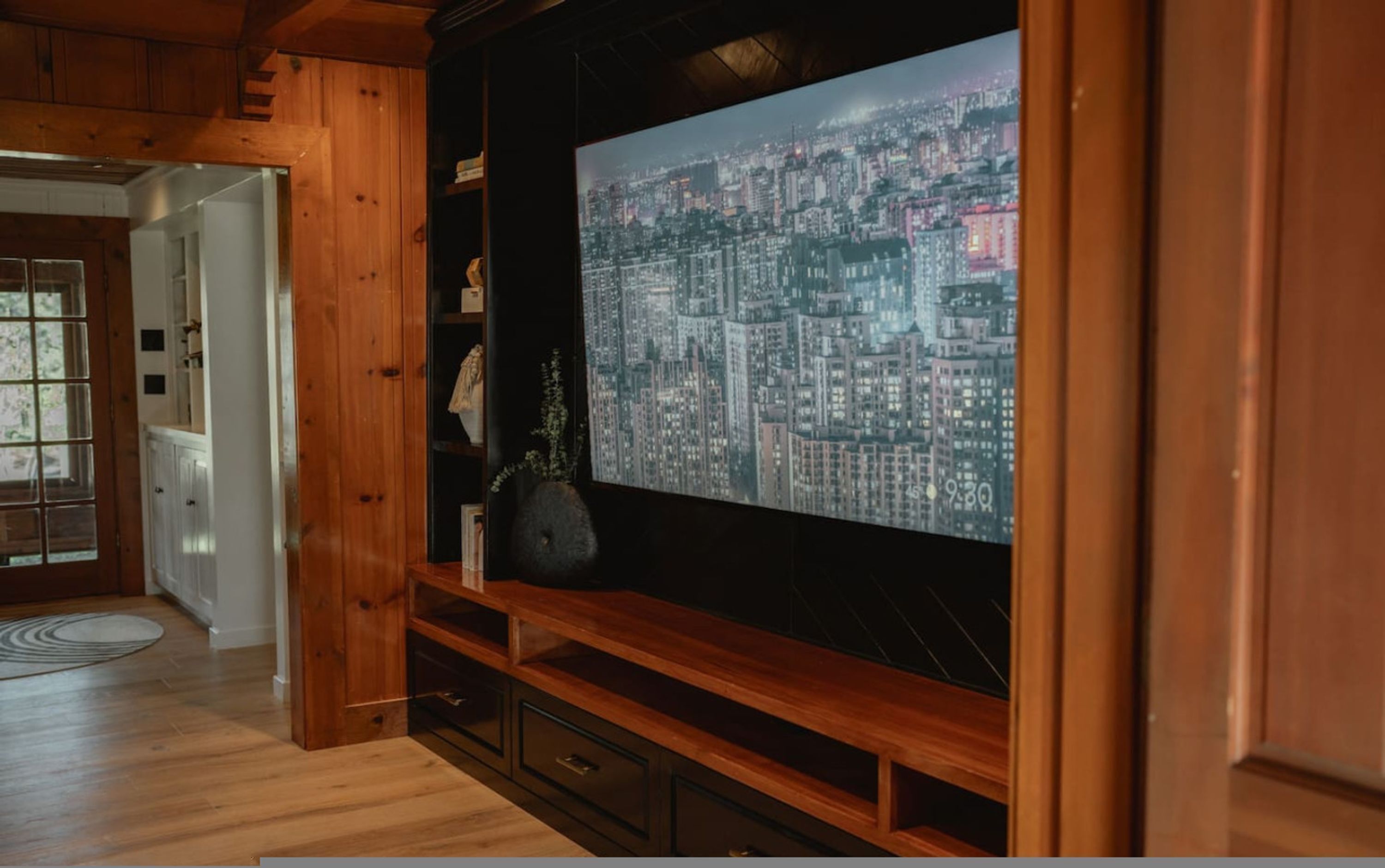 A living room with a large flat-screen TV mounted on a wooden wall, a wooden cabinet below, and a view of a cityscape at night.