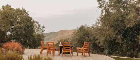 An outdoor seating area with wooden chairs and a table, surrounded by trees and a hilly landscape.