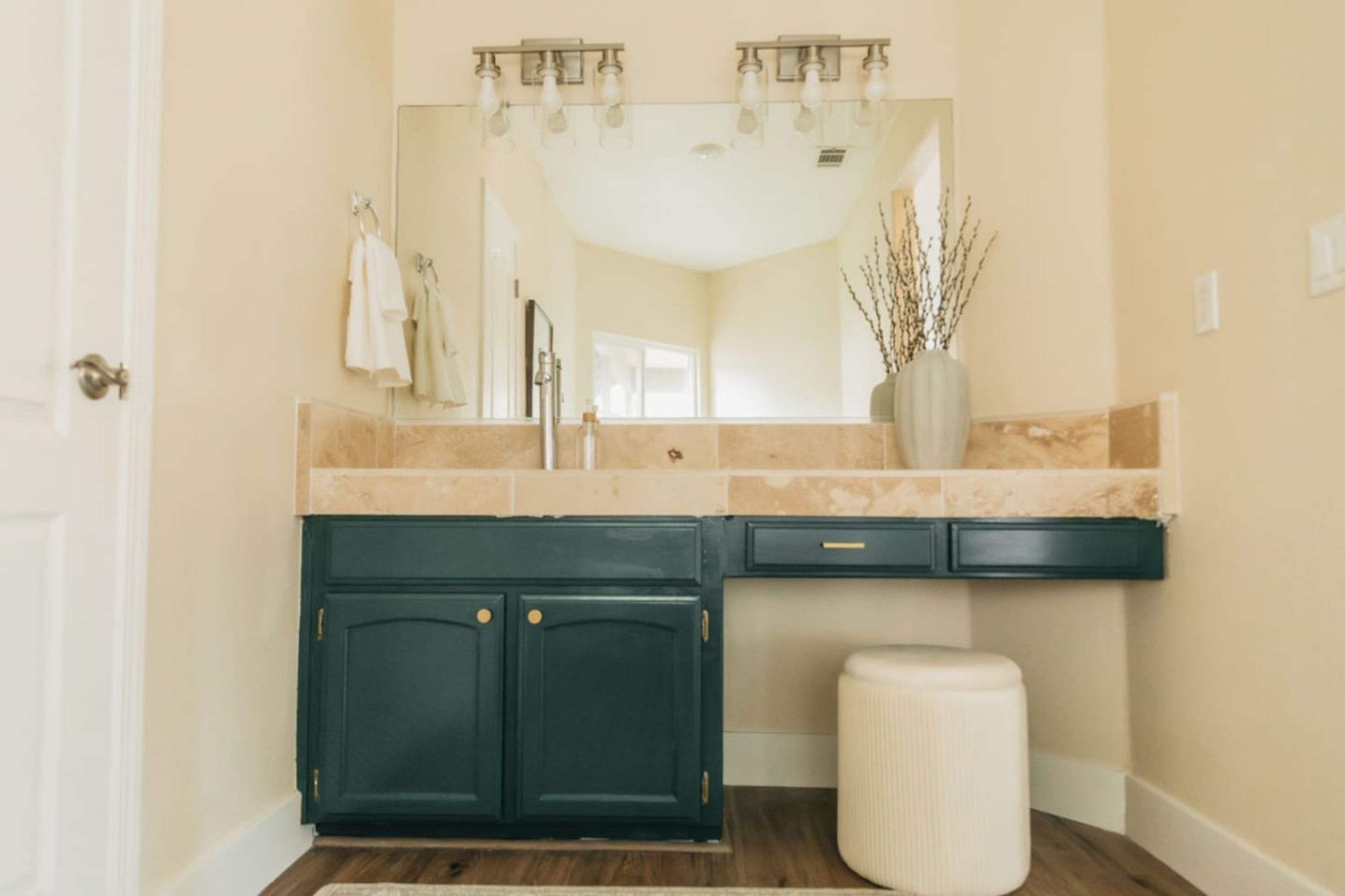 A bathroom with a beige tiled wall, a dark green vanity with two drawers, a white stool, and a mirror above the sink.