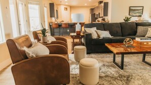 A modern living room with a dark sofa, leather armchairs, and a wooden coffee table. A kitchen is visible in the background with bar stools.