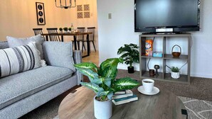 A living room with a grey sofa, a coffee table with books and a plant, a flat-screen TV on a stand, and a dining area with a chandelier. - Prime Location Yosemite Expedition by Casa Oso (Oakhurst)