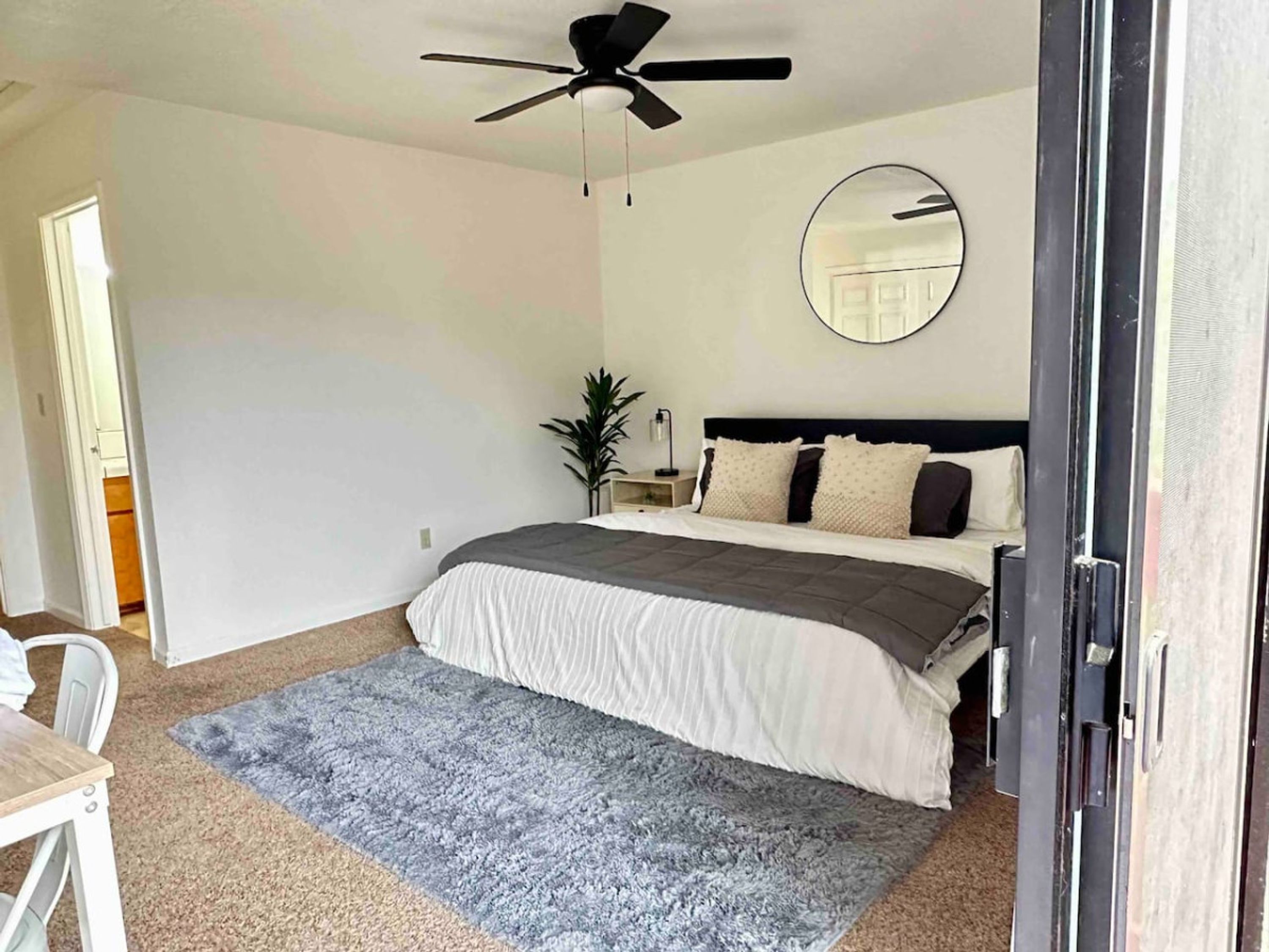 A bedroom with a bed, a ceiling fan, a bedside table with a lamp, a mirror on the wall, and a door leading to another room.