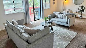 A modern living room with a grey sofa, a smaller grey armchair, a round coffee table, and a patterned rug. There is a sliding glass door leading to an outdoor area with a red deck. - Prime Location Yosemite Expedition by Casa Oso (Oakhurst)