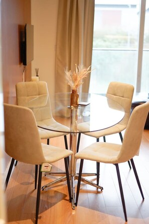 A modern dining area with a glass table surrounded by four chairs, a vase with dried plants, and a speaker mounted on the wall.
