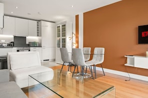 A modern living space with a glass dining table, white sofas, and a kitchen area with white cabinets and appliances.