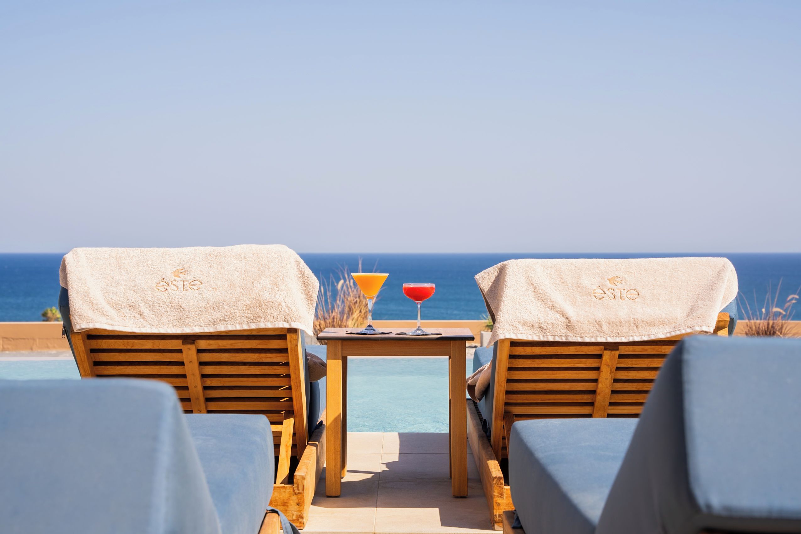 Two wooden lounge chairs with towels, a small table with a drink, and a view of the ocean.