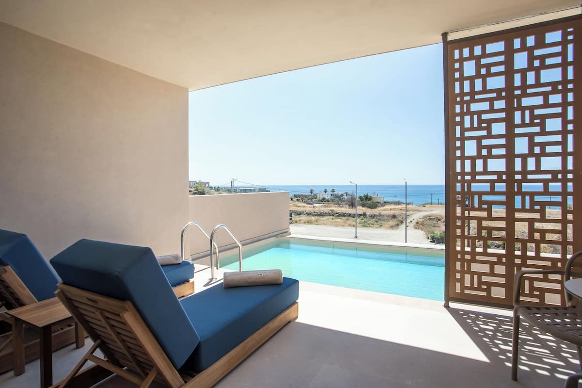 Un espace aménagé au bord de la piscine avec vue sur la mer, une chaise en bois, un fauteuil de relaxation bleu et une piscine dont l’eau reflète un ciel bleu clair.