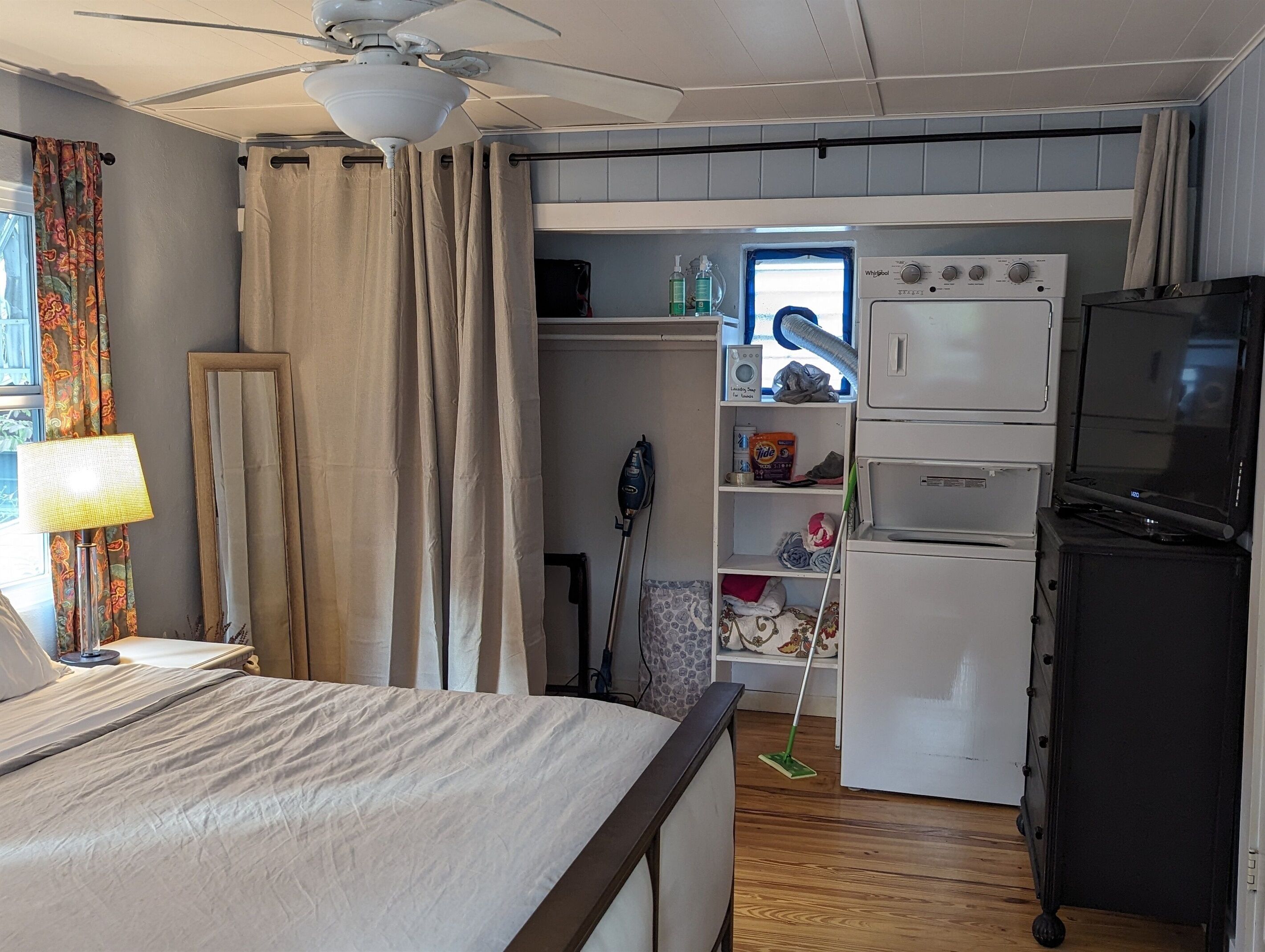 A compact hotel room with a bed, a ceiling fan, a TV, a wardrobe, and a small kitchenette.