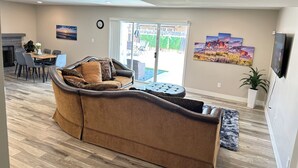 A living room with a sofa, a dining table with chairs, a fireplace, and a large window with a view of the outdoors. - A Getaway Home with a pool table, table tennis & a Pool. (Phoenix)
