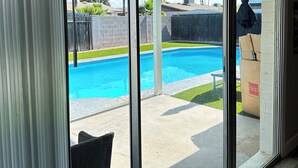 A view through a sliding glass door of a backyard with a swimming pool, a patio area, and a covered seating area. - A Getaway Home with a pool table, table tennis & a Pool. (Phoenix)