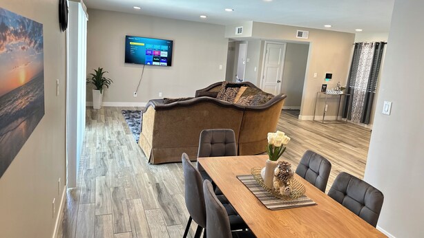 A modern living room with a flat-screen TV mounted on the wall, a cozy sectional sofa, and a dining area with a wooden table and chairs. - A Getaway Home with a pool table, table tennis & a Pool. (Phoenix)