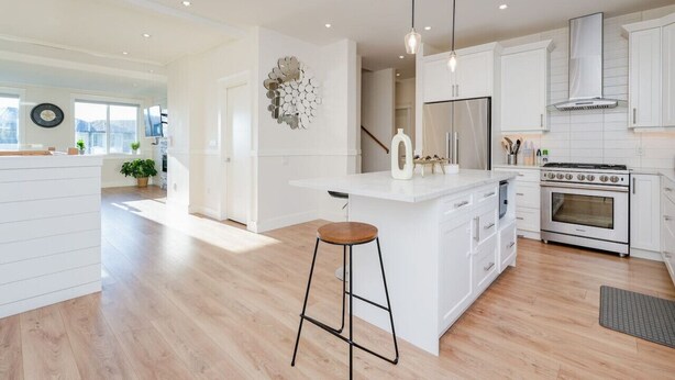 A modern kitchen with white cabinetry, a central island with a bar stool, a stove, and pendant lighting. - Elegant Home In Victoria BC (Langford)
