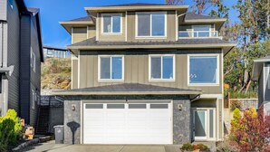 A two-story residential building with a garage, a stone foundation, and a hillside backdrop. - Elegant Home In Victoria BC (Langford)