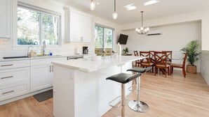 A modern kitchen with white cabinetry, a central island with bar stools, a dining area with wooden chairs, and pendant lighting. - Elegant Home In Victoria BC (Langford)