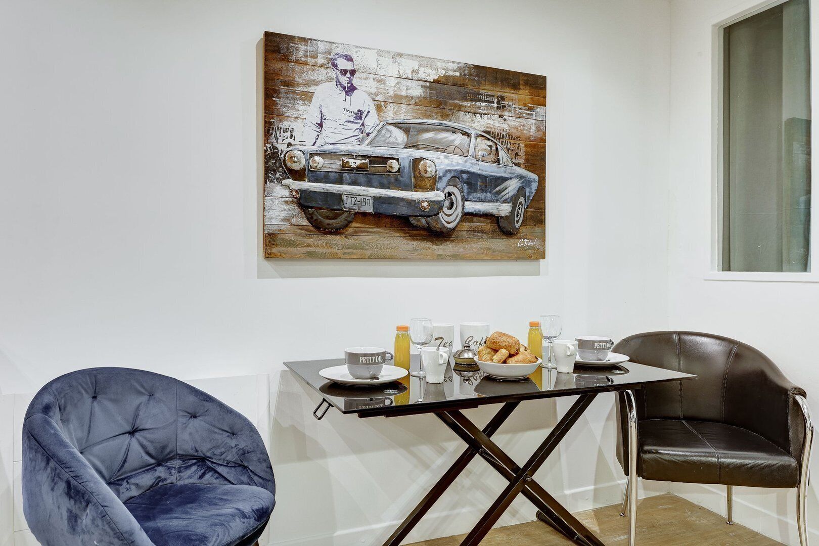 A dining area with a table set for breakfast, a painting of a vintage car, and two chairs.