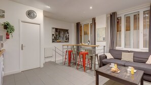 Una sala moderna con un sofá, una mesa de centro con cosas para el desayuno, una zona de comedor con taburetes, un reloj y una pintura de un paisaje urbano.