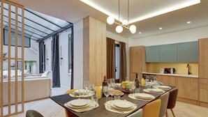 A modern dining area with a long table set for a meal, a kitchen in the background, and a glass partition separating the dining area from a living space. - TBS 174 - Exceptional flat in the heart of Paris (Paris)