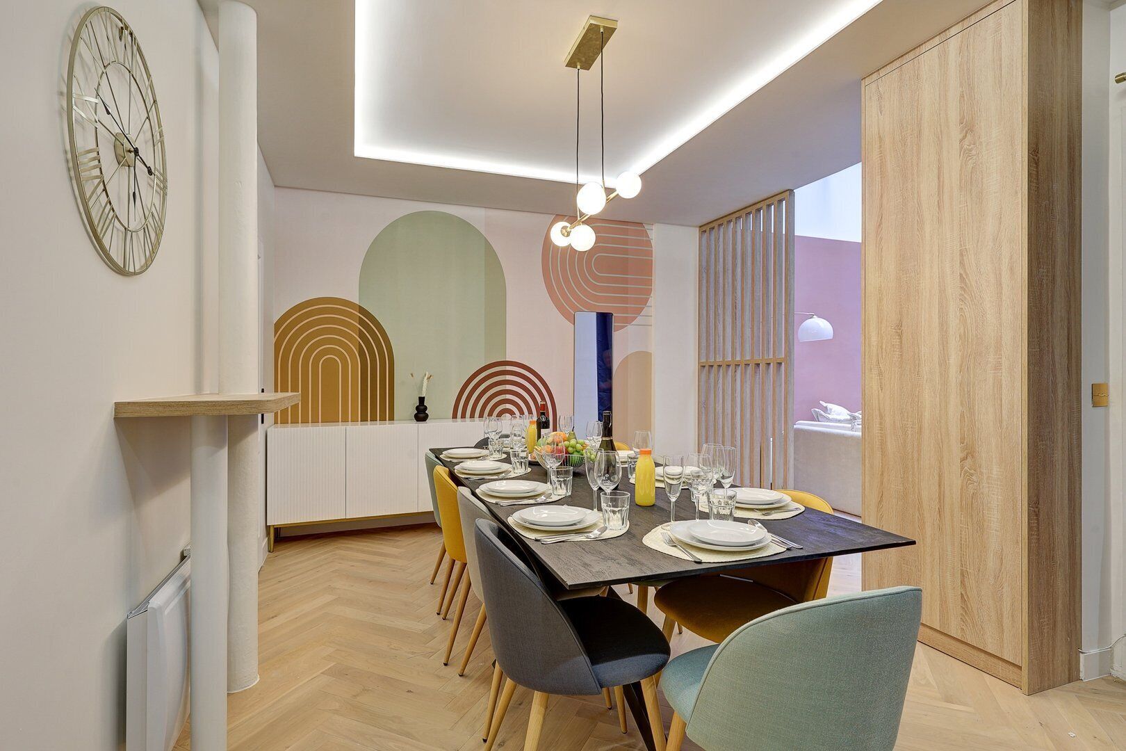 A dining area with a long table set for six, surrounded by chairs. A large wall clock is visible on the left.