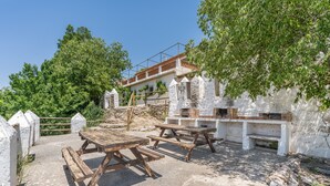 Un área de estar al aire libre rústica con bancos y mesas de madera, rodeada de muros de piedra blanca y vegetación.