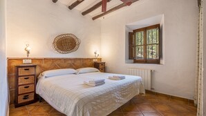 Un dormitorio con cabecera de madera, cama con ropa blanca, dos mesitas de noche con lámparas, un adorno de mimbre en la pared y una ventana con vistas.