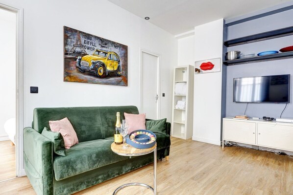Uma sala de estar com um sofá verde, um quadro de um carro amarelo, uma televisão e uma estante com louças.