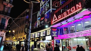 A brightly lit street with a prominent Ariston sign, indicating a commercial area with shops and outdoor seating.