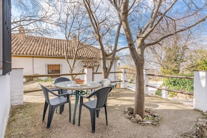 Eine Terrasse mit rundem Tisch und Stühlen, ein Baum und im Hintergrund ein Gebäude mit Ziegeldach.